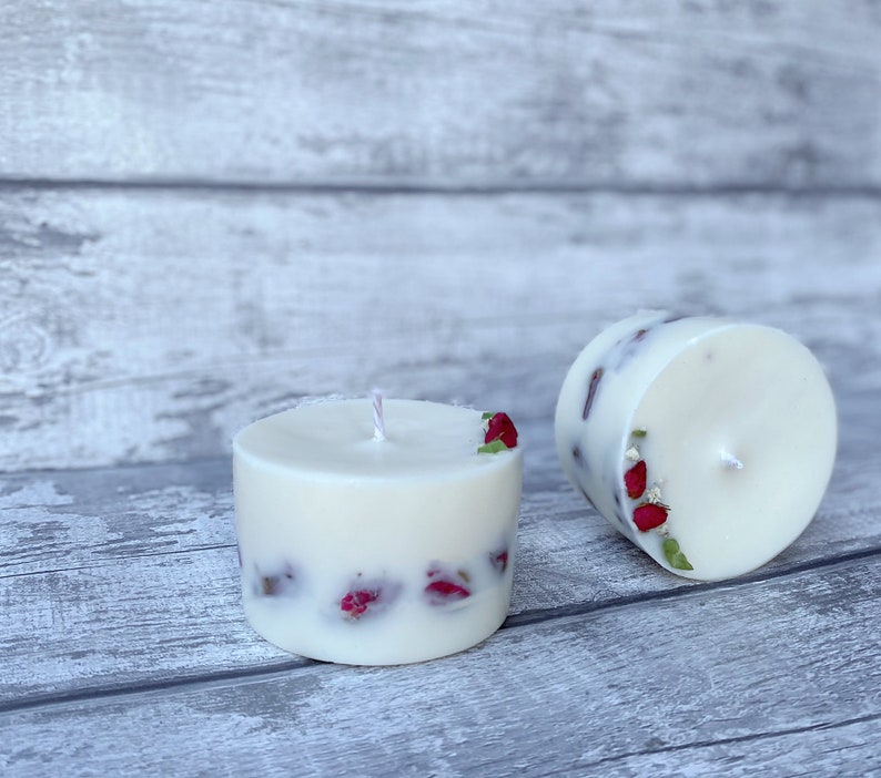May include: Two white pillar candles with dried red flowers embedded in the wax. The candles are on a light gray wooden surface.