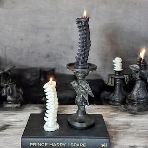 Pode incluir: Duas velas em forma de coluna vertebral, preta e branca, em casti&ccedil;ais pretos. Os casti&ccedil;ais s&atilde;o decorados com uma caveira e um corvo. As velas est&atilde;o sobre um livro preto com o texto "Prince Harry Spare" na capa.