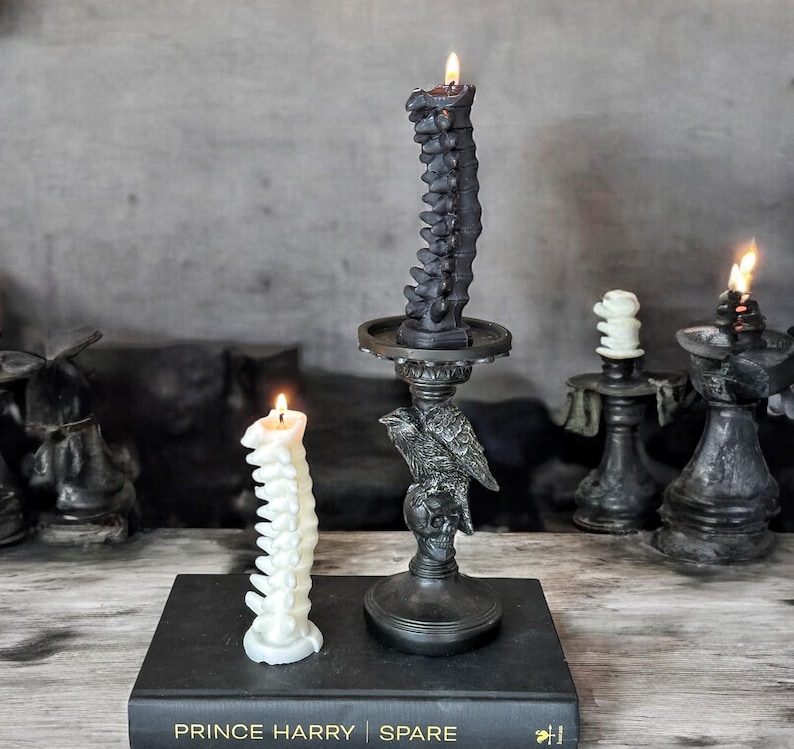 Pode incluir: Duas velas em forma de coluna vertebral, preta e branca, em casti&ccedil;ais pretos. Os casti&ccedil;ais s&atilde;o decorados com uma caveira e um corvo. As velas est&atilde;o sobre um livro preto com o texto "Prince Harry Spare" na capa.