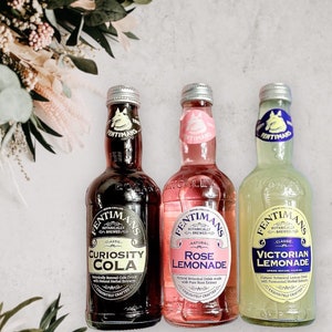 May include: Three glass bottles of Fentimans Botanically Brewed beverages. From left to right: Curiosity Cola, Rose Lemonade, and Victorian Lemonade. Each bottle has a unique label design and colour scheme.