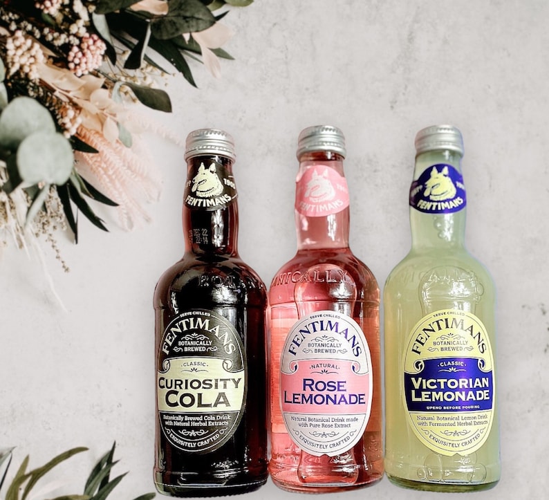 May include: Three glass bottles of Fentimans Botanically Brewed beverages. From left to right: Curiosity Cola, Rose Lemonade, and Victorian Lemonade. Each bottle has a unique label design and colour scheme.