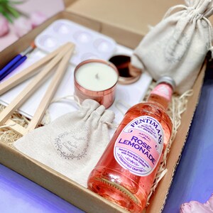 May include: A gift box filled with items including a small easel, a white paint palette, a candle in a copper container, a small hessian bag with a label that says "Alonix Candles", a larger hessian bag, and a bottle of Fentimans Rose Lemonade. The bottle has a pink label with the text "Fentimans Botanically Brewed Natural Rose Lemonade" and "Exquisitely Crafted".