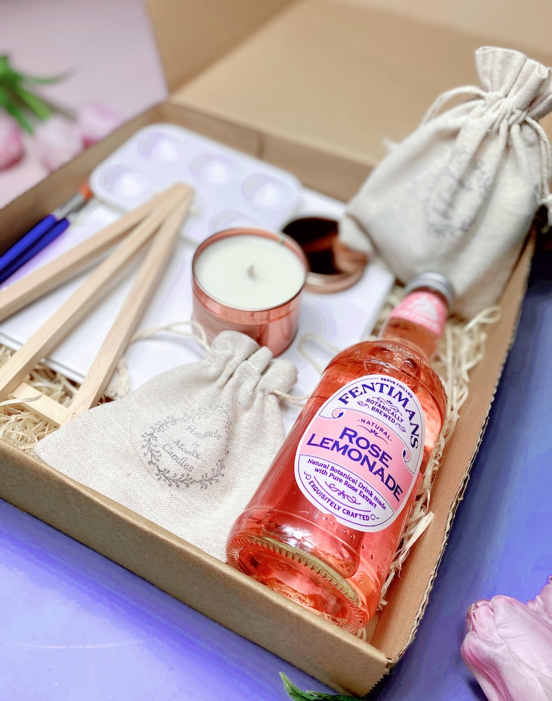 May include: A gift box filled with items including a small easel, a white paint palette, a candle in a copper container, a small hessian bag with a label that says "Alonix Candles", a larger hessian bag, and a bottle of Fentimans Rose Lemonade. The bottle has a pink label with the text "Fentimans Botanically Brewed Natural Rose Lemonade" and "Exquisitely Crafted".