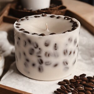Pu&ograve; includere: Una candela bianca con un design a forma di chicchi di caff&egrave; marroni. La candela &egrave; su una superficie bianca con chicchi di caff&egrave; sparsi intorno.