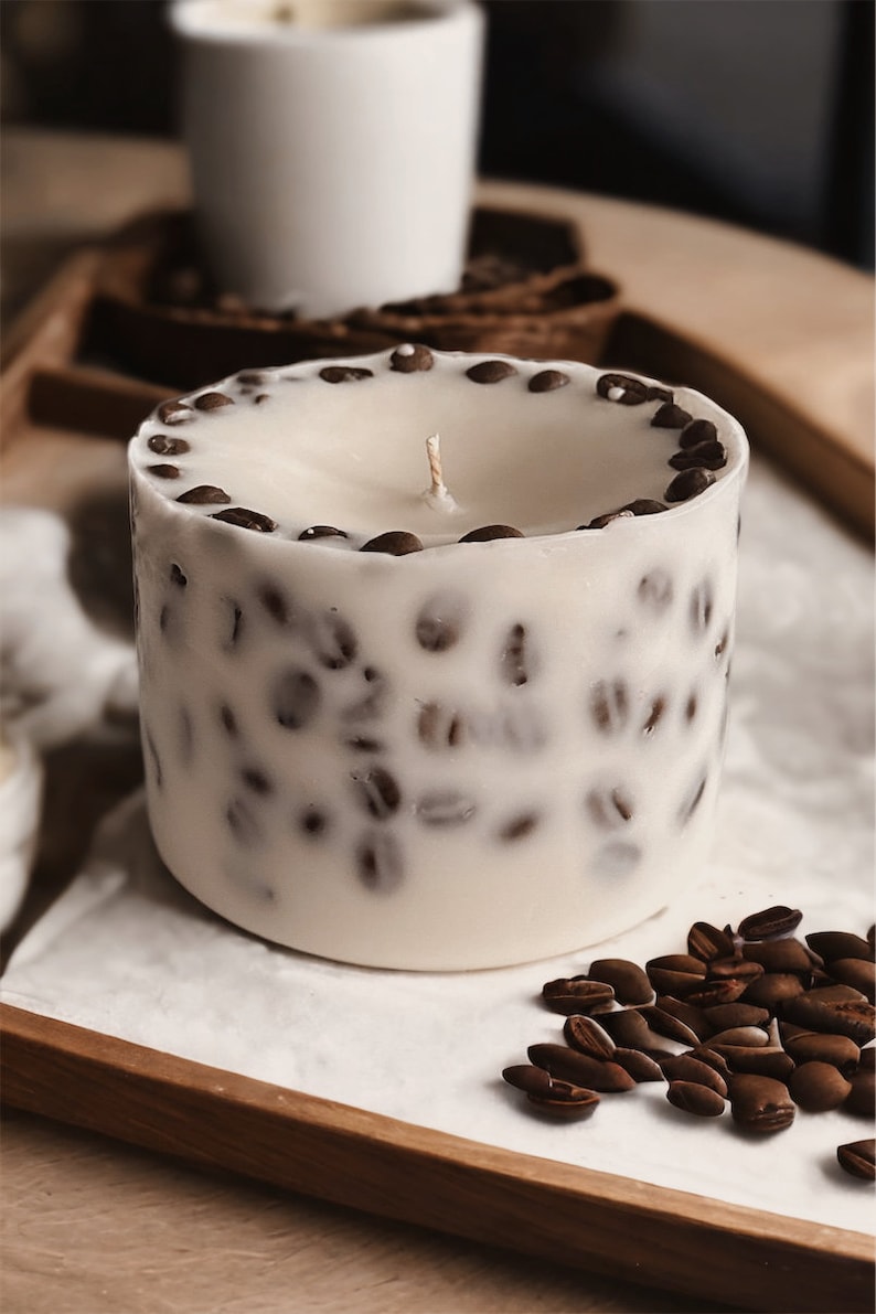 Pu&ograve; includere: Una candela bianca con un design a forma di chicchi di caff&egrave; marroni. La candela &egrave; su una superficie bianca con chicchi di caff&egrave; sparsi intorno.