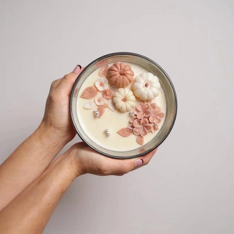 May include: A decorative candle in a clear glass container, featuring a cream-colored wax base. The candle is adorned with detailed, light-colored pumpkins, flowers, and leaves. The arrangement includes shades of pink, peach, and white, creating a seasonal, autumnal theme.