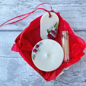 May include: A white gift box with red tissue paper. The box contains a white candle with dried flowers, a white wax melt with dried flowers, and a small glass vial with matches. The vial has a label that says "specially for you".