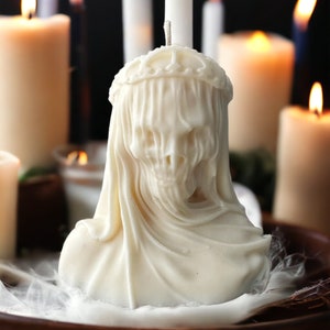 May include: White candle sculpted in the shape of a skull wearing a veil. The candle is surrounded by other white candles.