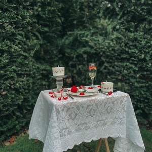 May include: A white lace tablecloth covers a wooden table set for two. Two white candles with red accents are on the table, along with a glass of champagne and a small gift box.