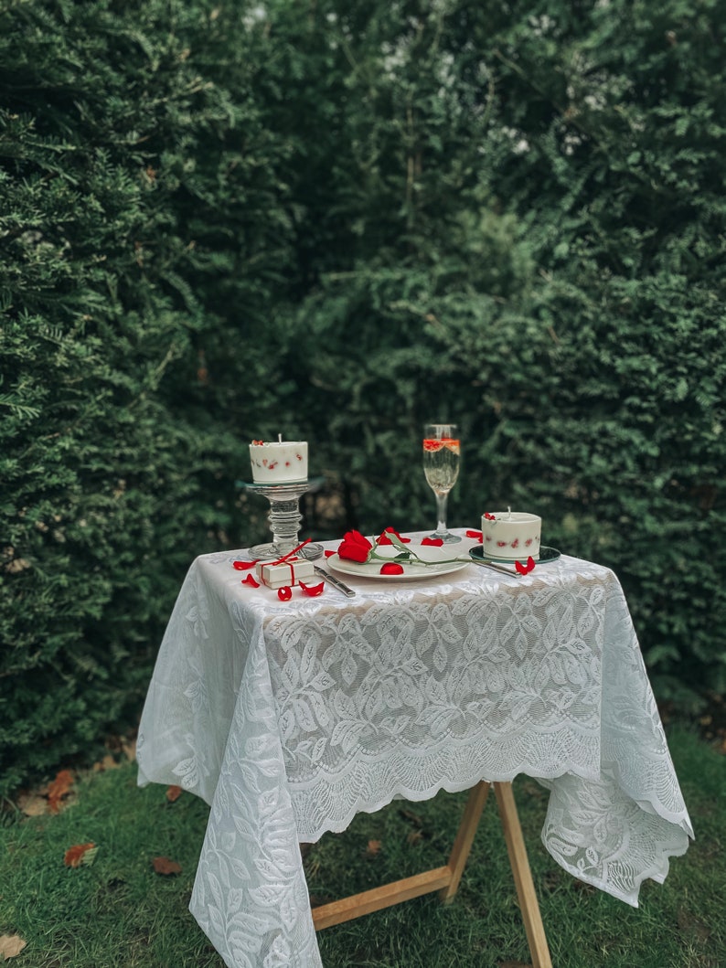 May include: A white lace tablecloth covers a wooden table set for two. Two white candles with red accents are on the table, along with a glass of champagne and a small gift box.