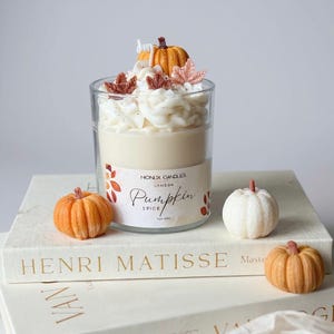 Op de afbeelding: Een geurkaars met pompoenkruiden in een glazen pot, versierd met een pompoen en bladeren, bekroond met witte glazuur. Het etiket luidt "Pumpkin Spice". De kaars wordt gepresenteerd met kleine pompoenversieringen.