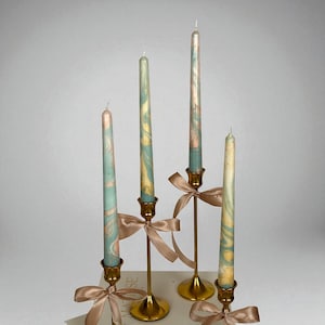 May include: Four taper candles with a marbled design in shades of blue, pink, and yellow. The candles are in gold-coloured candle holders, each adorned with a satin ribbon. The candles vary in height, creating a decorative display.