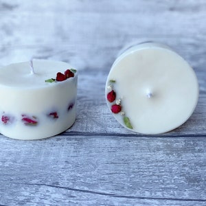 May include: Two white pillar candles with dried red rose petals embedded in the wax. One candle is lit and the other is unlit.
