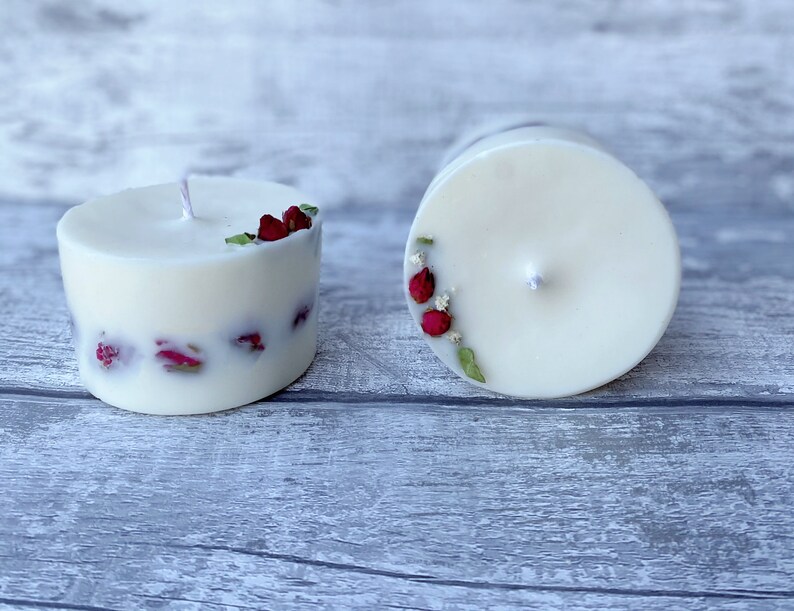 May include: Two white pillar candles with dried red rose petals embedded in the wax. One candle is lit and the other is unlit.