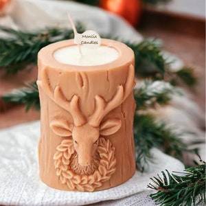 May include: A tan-colored candle shaped like a tree stump, featuring a carved deer head with a wreath. The candle has a white interior and a wick. The label says "Monlix Candles". The candle sits on a white cloth.
