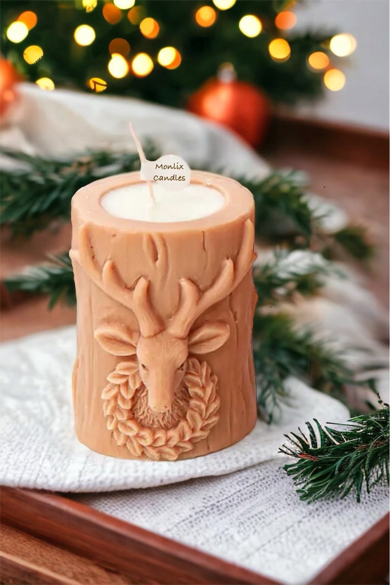 May include: A tan-colored candle shaped like a tree stump, featuring a carved deer head with a wreath. The candle has a white interior and a wick. The label says "Monlix Candles". The candle sits on a white cloth.
