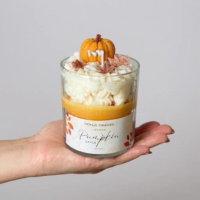 Puede incluir: Una vela en capas en un frasco de vidrio transparente, con un tema de especias de calabaza. La vela tiene una parte superior de cera en forma de calabaza, cera de color crema y una capa de cera naranja. La etiqueta dice "Pumpkin Spice" y "Monlux Candles London."