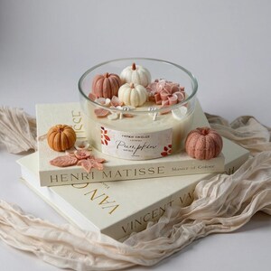 May include: A clear glass candle holder filled with beige wax, decorated with miniature pumpkins and floral accents. The candle sits atop two books, one labeled "Henri Matisse" and the other "Vincent Van Gogh". A small, orange pumpkin is placed next to the candle.