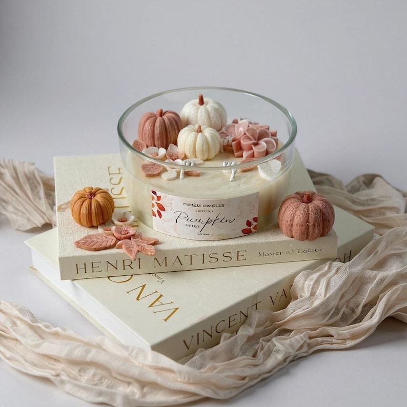 May include: A clear glass candle holder filled with beige wax, decorated with miniature pumpkins and floral accents. The candle sits atop two books, one labeled "Henri Matisse" and the other "Vincent Van Gogh". A small, orange pumpkin is placed next to the candle.