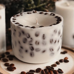 Pu&ograve; includere: Una candela bianca con un designo a forma di chicchi di caff&egrave; marroni. La candela &egrave; appoggiata su un sottobicchiere di legno con chicchi di caff&egrave; sparsi intorno.