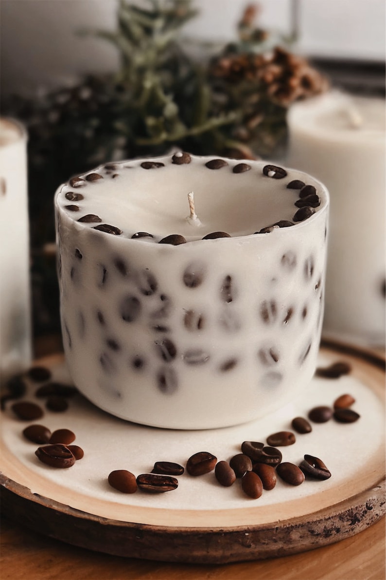 Pu&ograve; includere: Una candela bianca con un designo a forma di chicchi di caff&egrave; marroni. La candela &egrave; appoggiata su un sottobicchiere di legno con chicchi di caff&egrave; sparsi intorno.