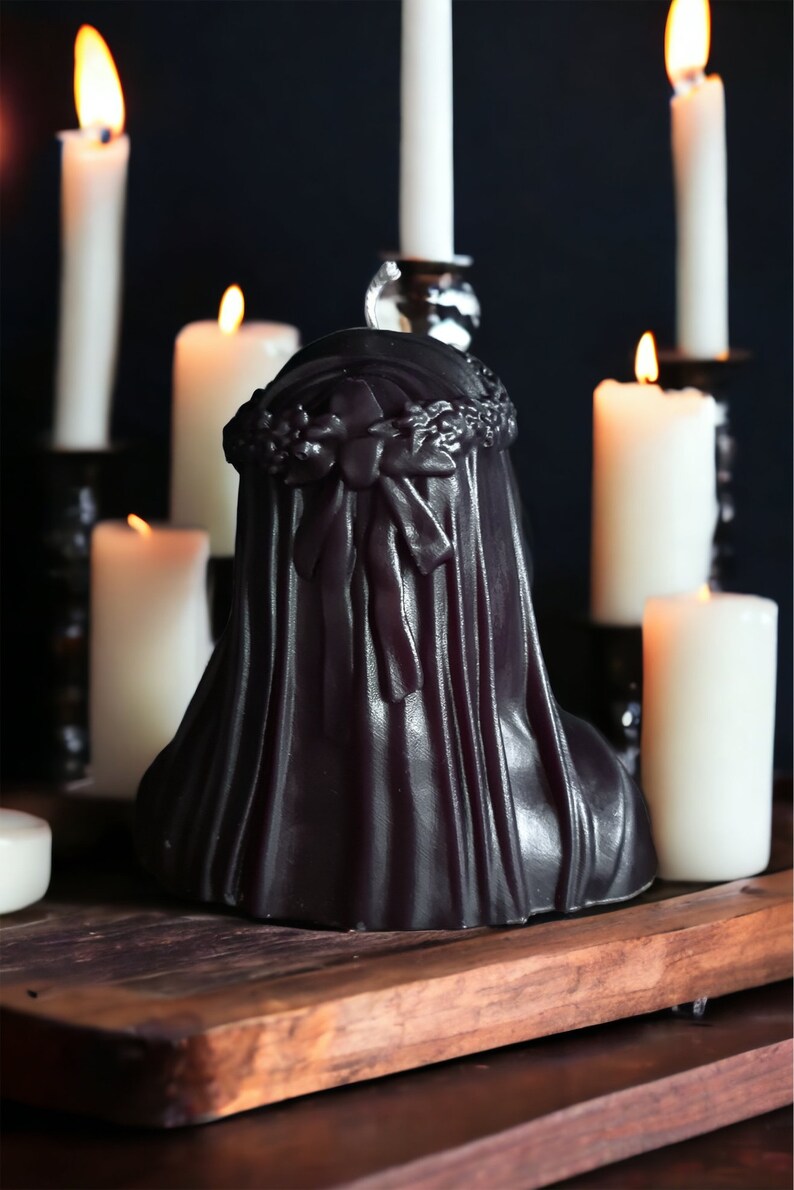 May include: A black candle in the shape of a woman wearing a veil and a floral crown. The candle is surrounded by white candles on a wooden surface.