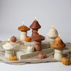 May include: A selection of mushroom-shaped candles in shades of brown and cream, with lit wicks. Small pumpkin decorations in orange, white, and brown are arranged around the candles, creating an autumnal display.
