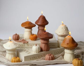 May include: A selection of mushroom-shaped candles in shades of brown and cream, with lit wicks. Small pumpkin decorations in orange, white, and brown are arranged around the candles, creating an autumnal display.