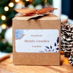 May include: A brown cardboard box with a label that reads "Handmade Monlix Candles London." The box is tied with a brown ribbon and sits on a wooden tray. The background features blurred Christmas lights and a pine cone.