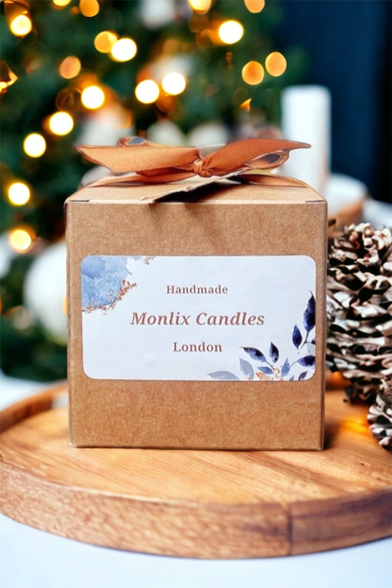 May include: A brown cardboard box with a label that reads "Handmade Monlix Candles London." The box is tied with a brown ribbon and sits on a wooden tray. The background features blurred Christmas lights and a pine cone.