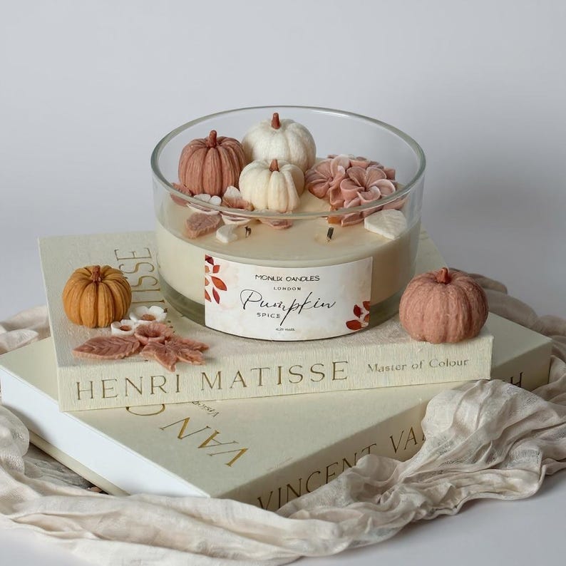 May include: A large glass candle with a pumpkin spice scent, decorated with small pumpkins and flowers. The candle sits atop stacked books with the titles "Henri Matisse" and "Vincent Van Gogh."