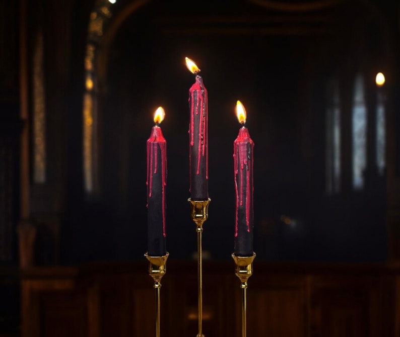 May include: Three black candles with red wax dripping down the sides are lit and placed on a gold candelabra.