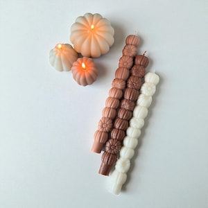 Può includere: Una collezione di candele a tema autunnale in varie forme e colori. L'immagine presenta tre candele a forma di zucca nei toni del pesca e del bianco, con stoppini accesi. Sono presenti anche diverse candele lunghe impilate nei toni del marrone e del bianco.