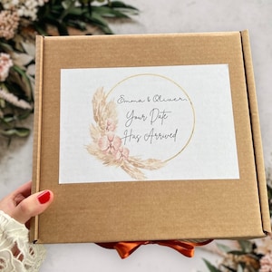 May include: A brown cardboard box with a white label. The label has a gold circle with a floral design and the text "Emma & Oliver, Your Date Has Arrived".