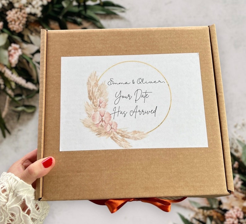 May include: A brown cardboard box with a white label. The label has a gold circle with a floral design and the text "Emma & Oliver, Your Date Has Arrived".