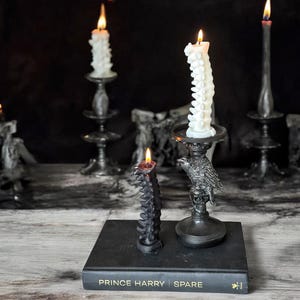 Pode incluir: Duas velas pretas em forma de coluna vertebral, uma acesa, sobre um livro preto com o t&iacute;tulo "Prince Harry Spare" na capa. As velas est&atilde;o sobre um casti&ccedil;al de metal preto com um design de corvo.