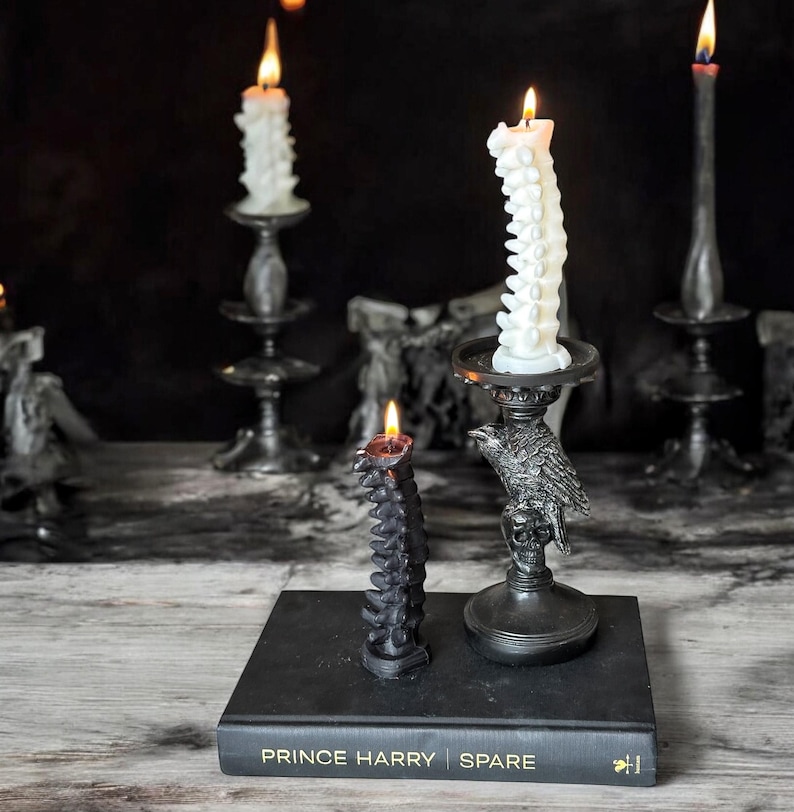 Pode incluir: Duas velas pretas em forma de coluna vertebral, uma acesa, sobre um livro preto com o t&iacute;tulo "Prince Harry Spare" na capa. As velas est&atilde;o sobre um casti&ccedil;al de metal preto com um design de corvo.