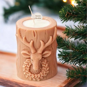 May include: A tan-colored candle shaped like a tree stump, featuring a detailed deer head with antlers and a wreath. The candle has a white wax interior and a wick. The text "Monlix Candles" is on a small tag. The candle sits on a wooden tray.