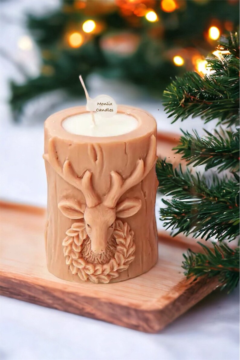 May include: A tan-colored candle shaped like a tree stump, featuring a detailed deer head with antlers and a wreath. The candle has a white wax interior and a wick. The text "Monlix Candles" is on a small tag. The candle sits on a wooden tray.