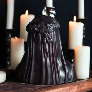 May include: A black candle in the shape of a woman wearing a veil and a flower crown. The candle is surrounded by white candles on a wooden surface.