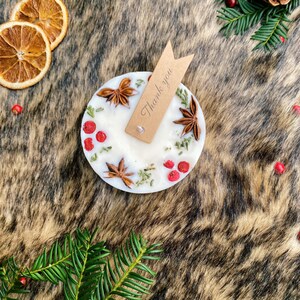 May include: A white candle with a natural, rustic design. The candle is decorated with dried herbs, star anise, and red berries. A small brown tag with the text "Thank you" is attached to the candle.