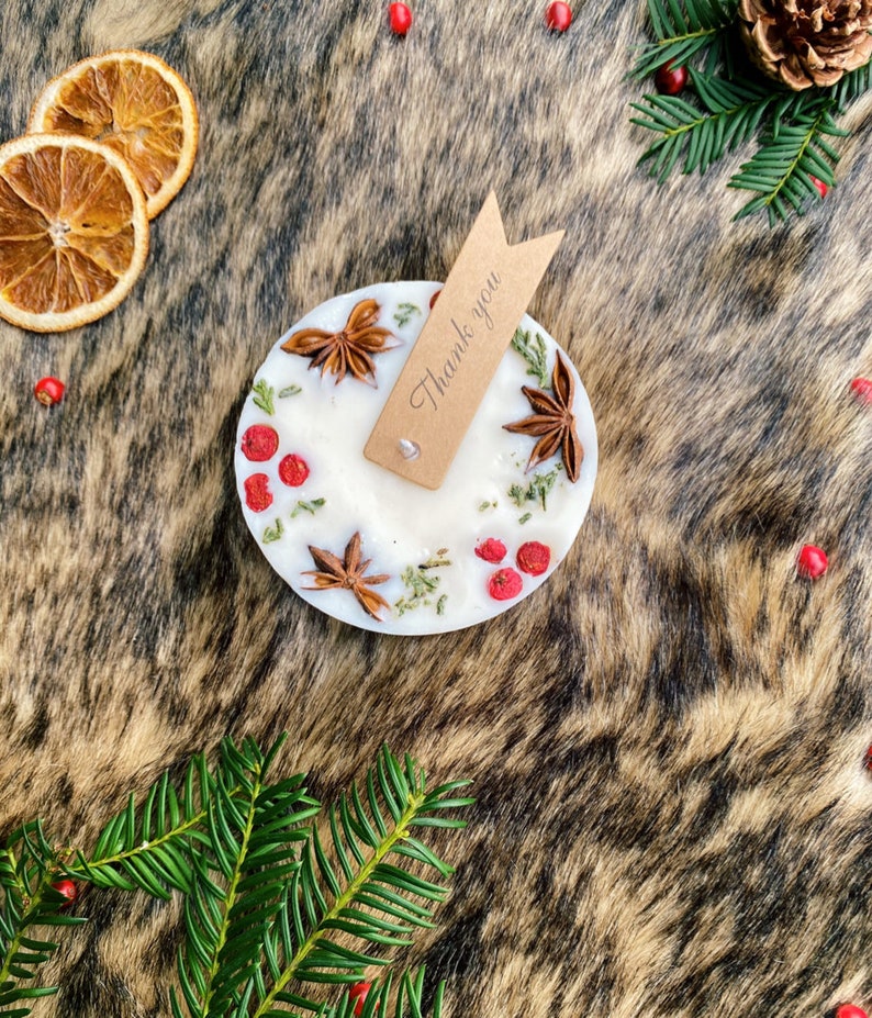 May include: A white candle with a natural, rustic design. The candle is decorated with dried herbs, star anise, and red berries. A small brown tag with the text "Thank you" is attached to the candle.