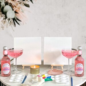 May include: Two white canvases on easels, two pink cocktails, two bottles of pink lemonade, a candle, paintbrushes, a paint palette, and a small white bag on a black and white table.