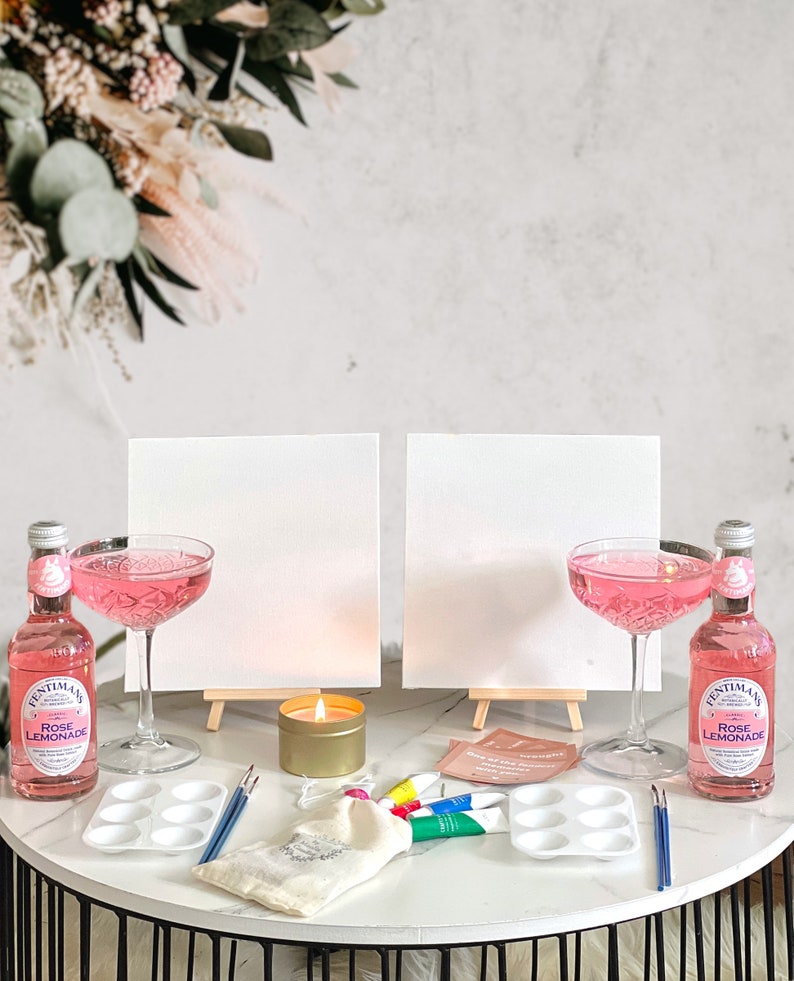 May include: Two white canvases on easels, two pink cocktails, two bottles of pink lemonade, a candle, paintbrushes, a paint palette, and a small white bag on a black and white table.