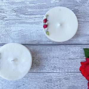 May include: Two white pillar candles with dried rose buds and greenery embedded in the wax. The candles are on a gray wooden surface. There are red roses with green leaves in the bottom right corner of the image.