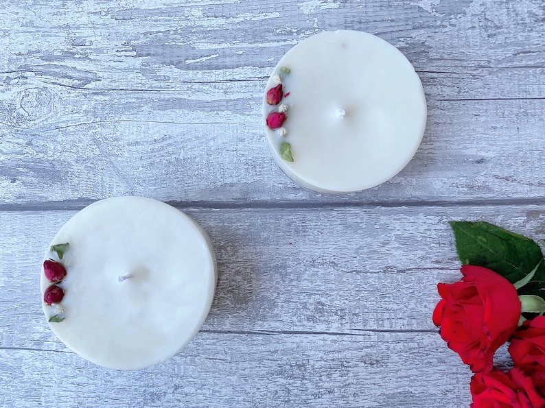 May include: Two white pillar candles with dried rose buds and greenery embedded in the wax. The candles are on a gray wooden surface. There are red roses with green leaves in the bottom right corner of the image.