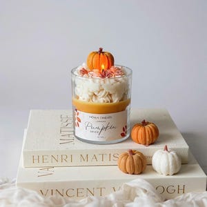 Puede incluir: Una vela de cristal transparente con aroma a especias de calabaza, coronada con una peque&ntilde;a calabaza y cera blanca. La vela est&aacute; sobre una pila de libros con los nombres Matisse, Henri Matisse y Vincent Van Gogh. Tres peque&ntilde;as calabazas est&aacute;n al lado de la vela.