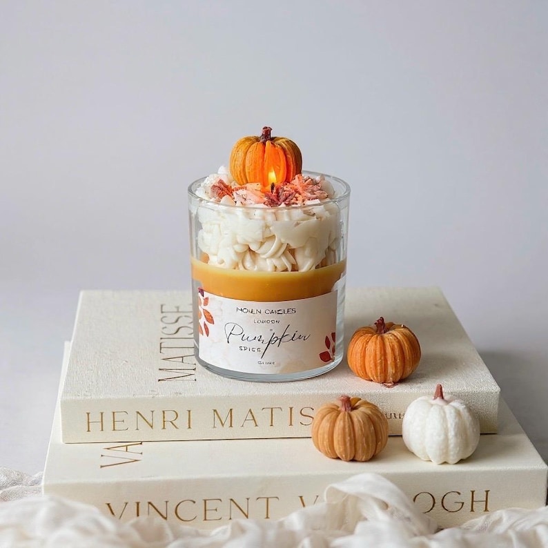Puede incluir: Una vela de cristal transparente con aroma a especias de calabaza, coronada con una peque&ntilde;a calabaza y cera blanca. La vela est&aacute; sobre una pila de libros con los nombres Matisse, Henri Matisse y Vincent Van Gogh. Tres peque&ntilde;as calabazas est&aacute;n al lado de la vela.
