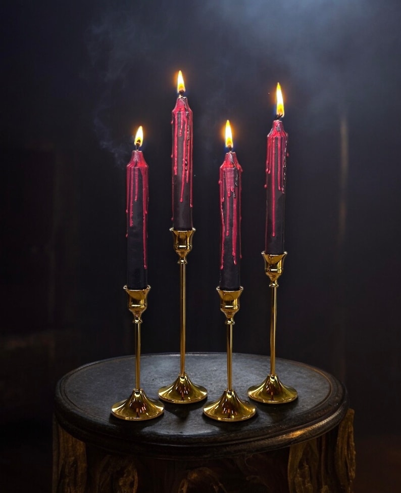 May include: Four black candles with red wax dripping down the sides are lit and placed on gold candle holders on a dark brown wooden surface.