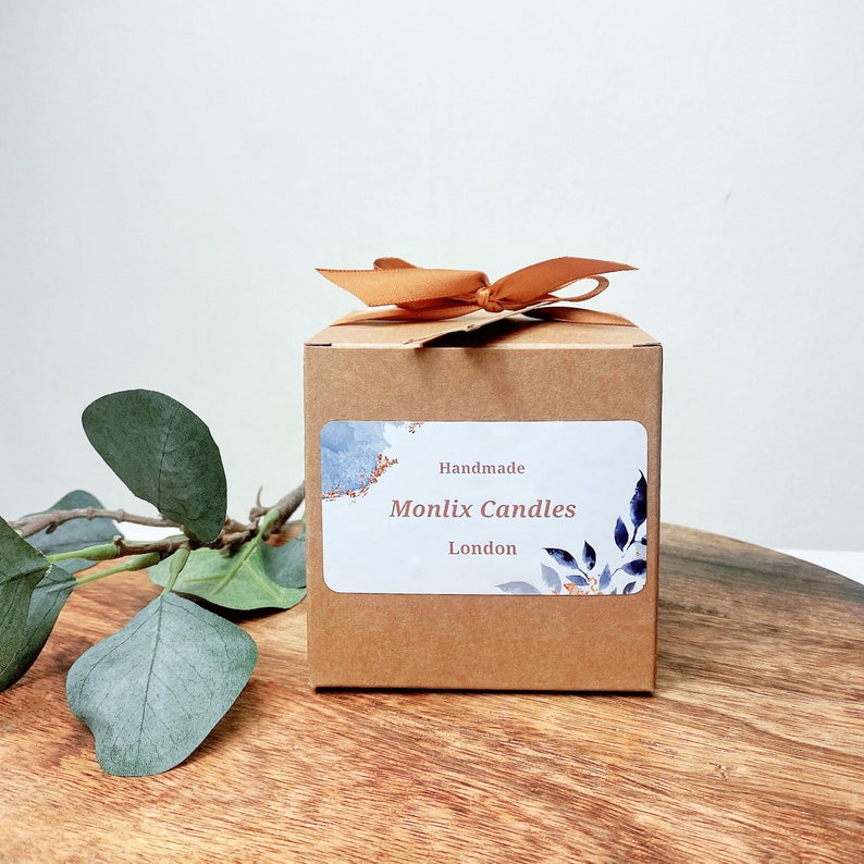 May include: A brown cardboard box with a gold ribbon tied around it. The box has a white label with the text "Handmade Monlix Candles London" and a blue and orange watercolor design.